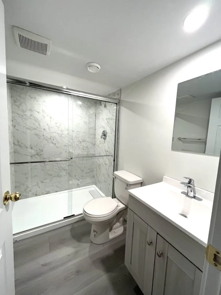 A basement bathroom with a marble-look shower surround and a grey vanity